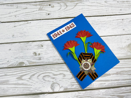 Victory Day greeting card with red paper carnations, St. George ribbon, and Soviet WWII medal on blue background. 1941 - 1945 date emphasizes memory of the war. Handmade craft on wooden surfaceの写真素材