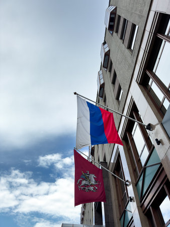 Russian flag and Moscow flag at entrance to building against cloudy sky. State symbol of Russia and Moscow region on facade of government. Vertical photo of flags of the country in flagpolesの写真素材