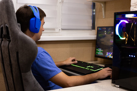 Young man wearing blue headphones, playing video game on desktop computer with RGB lights, gaming...