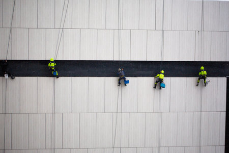 Industrial climbers hang at height on building exterior. Construction climbers are cleaning the system on building facade. Work at height in weatherの写真素材