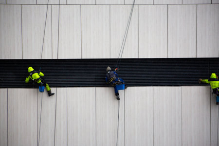 Industrial climber in protective clothing hangs from safety ropes while working on the side of building. Dangerous profession at height. The process of hard work in adverse weatherの写真素材