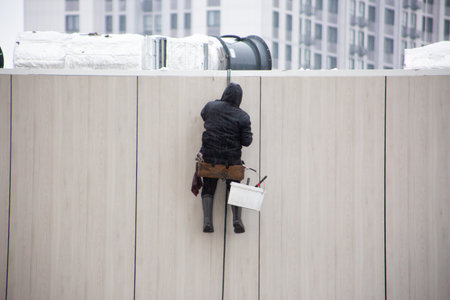 Industrial climber tests his safety ropes and carabiner for safety while working at height. Difficult work in challenging weather conditions. Man dangerous jobの写真素材