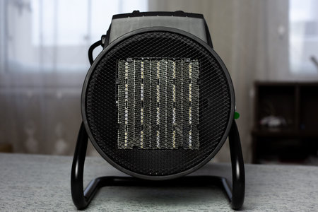 Room heater radiator behind protective mesh in round housing against backdrop of windows. Direct view of room heater radiator. Concept for increasing temperatureの写真素材