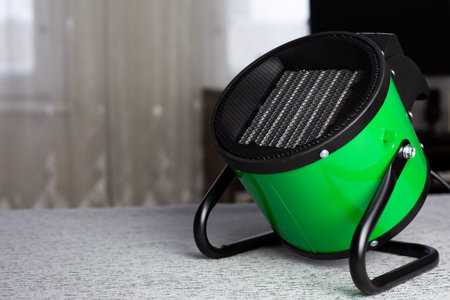 Raising the room temperature. Electric round heater with radiator and fan for circulating warm air throughout the room. Heating the room for living. Green heat gun on stand is pointed upwardsの写真素材