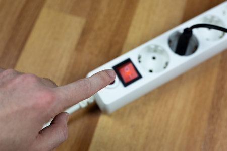 Person hand reaches towards red power switch on white electrical strip placed on wooden surface....