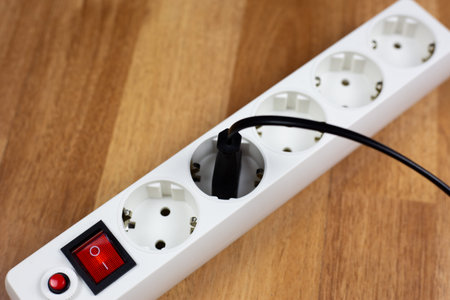 White surge protector with red switch and safety button rests on wood. Dark plug is engaged in one...