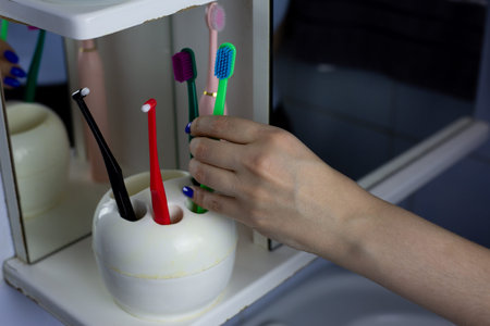 Woman hand picks up toothbrush from family set of toothbrushes in cup. Toothbrush holder sits on...