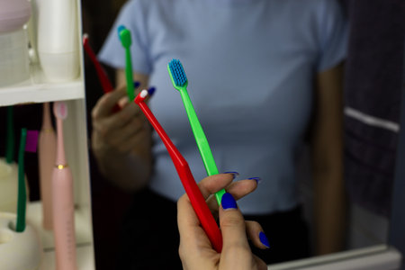 Woman stands in front of bathroom mirror holding red and green toothbrushes in hand. Scene shows daily activity of brushing teeth. Morning as person prepares for dayの写真素材