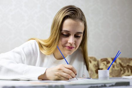 Young blonde smiles sincerely while painting her picture. Enjoyment and laughter come from favorite hobby. Creating drawing at home. Authentic emotions while creating funny pictureの写真素材