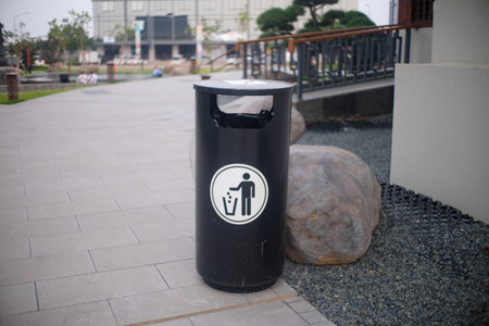 A sleek black trash bin with a simple waste disposal icon, placed on a tiled pathway next to decorative rocks in a public outdoor area.の写真素材