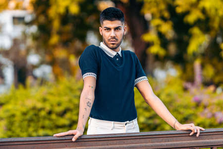 Portrait if a man with modern clothes and tattoos leaning on a rail in a parkの写真素材