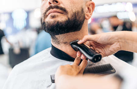 Hands grooming the beard of a man with an electric razor in a barber shop