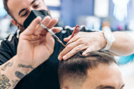 Barber with mask cutting the hair of a client using scissors in a barbers shopの写真素材