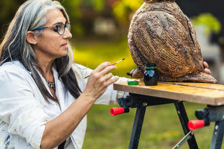 Woman with grey hair in gown painting a polystyrene sculpture in a garden