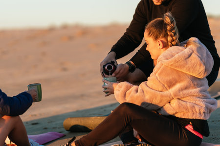 Man pouring tea on a cup to a women sitting on the beach after a yoga classの写真素材