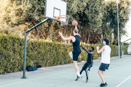 Tall man jumping to throw a ball into a basket while playing basketball with friendsの写真素材