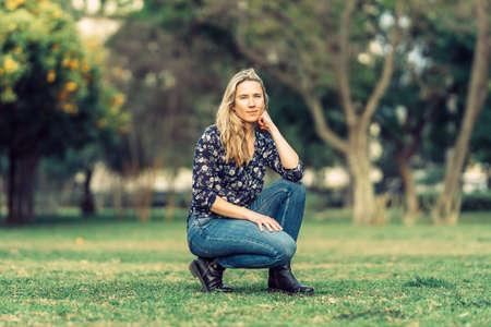 blonde-haired woman poses for the camera squatting in a park