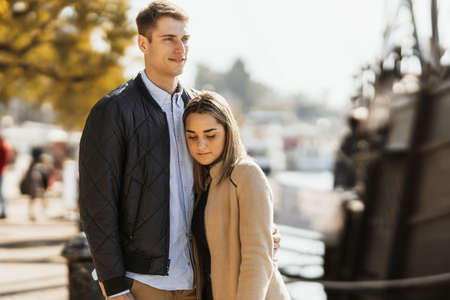 A young blonde girl rests her head on her boyfriends chest while looking down at the ground.