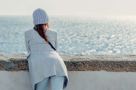 Rear view of a woman leaning against the promenade wall overlooking the blue ocean