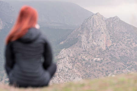 unfocused red-haired woman in the foreground looks up at the rocky mountain through the mist