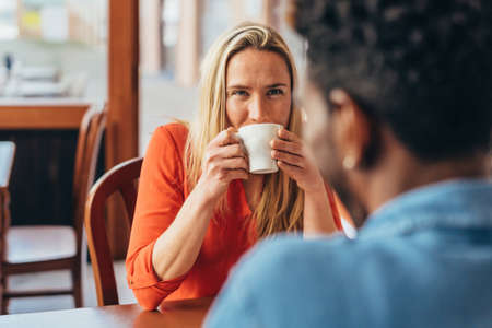 blonde woman looks attentively at black male companion while drinking coffee