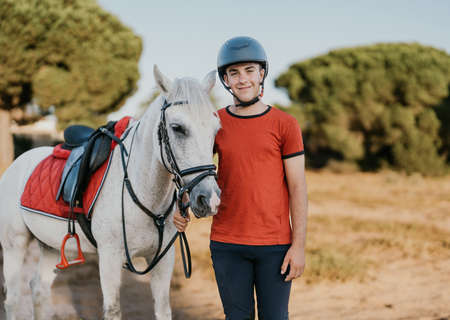 portrait of young smiling rider standing next to his white horse in a pine groveの写真素材