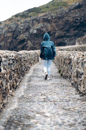 woman wearing a raincoat walking along a paved road enclosed by two stone wallsの写真素材