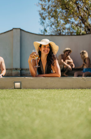 vertical photo of a smiling young woman wearing a pamela and leaning on the edge of a poolの写真素材