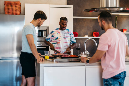 3 friends converse in a kitchen while one cuts meat with a knifeの写真素材