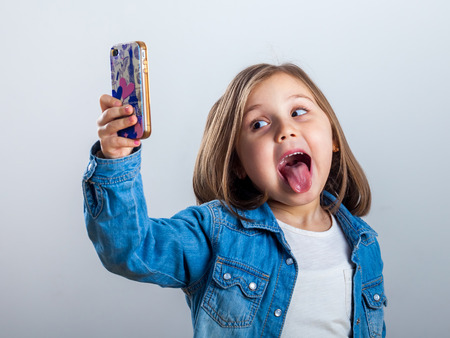 Beautiful little girl is doing selfie using a smart phone, on light background.