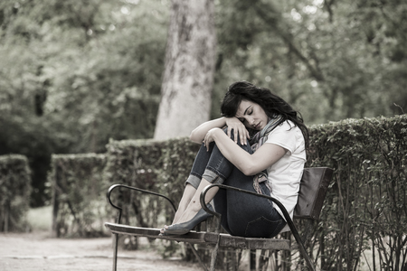 attractive beautiful latin woman sitting on a park bench feeling sad and depressed in a park outside in europeの写真素材