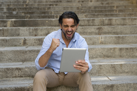 young attractive latin man sitting on city staircase working with digital tablet pad looking satisfied and confident in new business technology and lifestyle conceptの写真素材