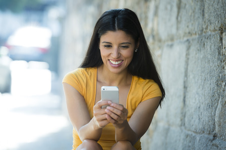Happy young latin woman talking and texting on smart phone, wearing casual clothes sitting in with a take away coffee in a urban bench の写真素材
