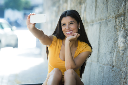 young attractive and sweet latin woman smiling happy taking self portrait selfie photo with mobile phone camera outdoors at urban city background in blogger conceptの写真素材