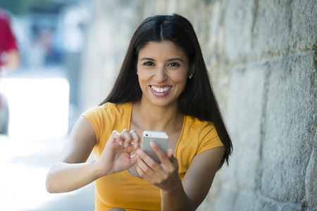Happy young latin woman talking and texting on smart phone, wearing casual clothes sitting in with a take away coffee in a urban bench の写真素材