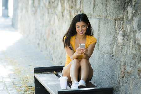 Happy young latin woman talking and texting on smart phone, wearing casual clothes sitting in with a take away coffee in a urban bench の写真素材