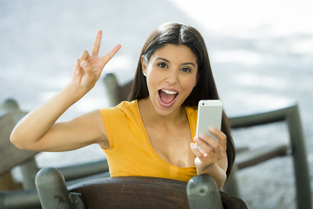 Happy young latin woman talking and texting on smart phone giving the peace sign, wearing casual clothes sitting in with a take away coffee in a urban bench 

の写真素材