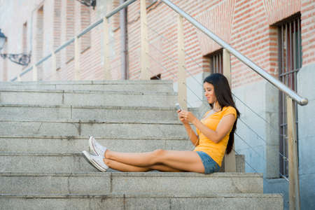 attractive latin woman in her twenties happy working on her mobile smart phone while drinking a coffee sitting on stairs. の写真素材