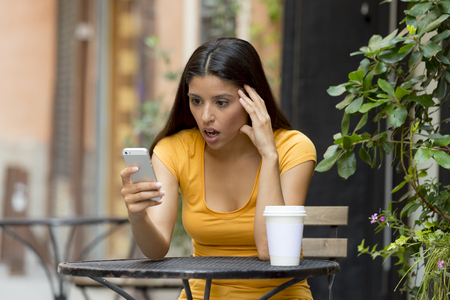 attractive latin woman in her twenties shocked while reading a tex message on her smart phone at a coffeeshopの写真素材