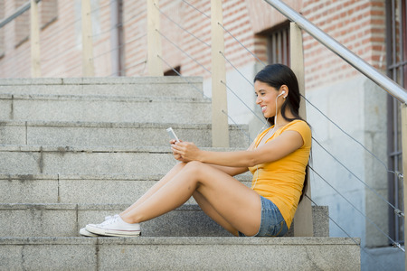 attractive latin woman in her twenties happy listening to music on her mobile smart phone while drinking a coffee sitting on stairs. の写真素材