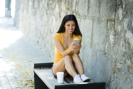 Happy young latin woman talking and texting on smart phone, wearing casual clothes sitting in with a take away coffee in a urban bench の写真素材