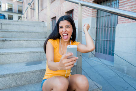 attractive latin woman in her twenties happy listening to music on her mobile smart phone while drinking a coffee sitting on stairs. の写真素材