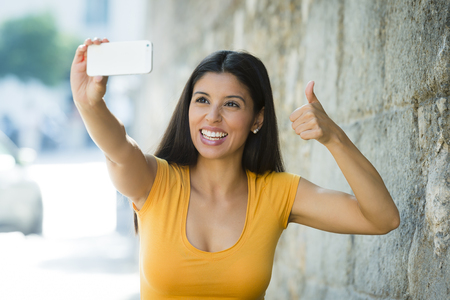 young attractive and sweet latin woman smiling happy taking self portrait selfie photo with mobile phone camera outdoors at urban city background in blogger conceptの写真素材