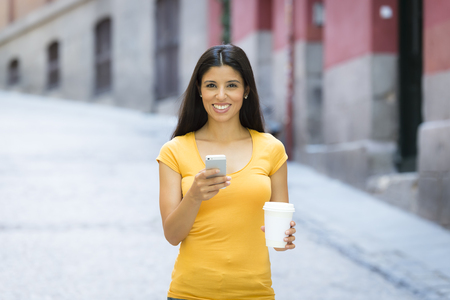 Happy young latin woman talking and texting on smart phone, wearing casual clothes sitting in with a take away coffee in a urban bench の写真素材
