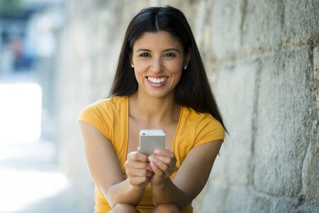 Happy young latin woman talking and texting on smart phone, wearing casual clothes sitting in with a take away coffee in a urban bench の写真素材