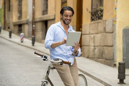 young happy attractive hispanic man in casual trendy clothes smiling cheerful using internet with digital tablet pad on vintage cool retro bicycle in modern stylish lifestyle urban style concept の写真素材