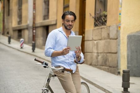 young happy attractive hispanic man in casual trendy clothes smiling cheerful using internet with digital tablet pad on vintage cool retro bicycle in modern stylish lifestyle urban style concept の写真素材