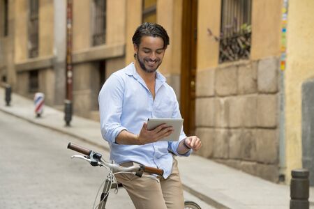 young happy attractive hispanic man in casual trendy clothes smiling cheerful using internet with digital tablet pad on vintage cool retro bicycle in modern stylish lifestyle urban style concept の写真素材