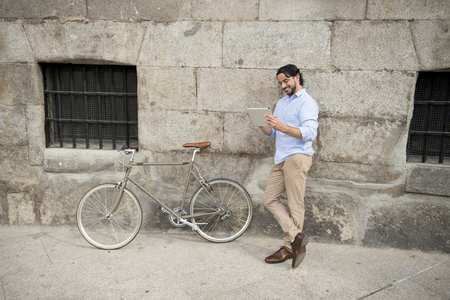 young happy attractive hispanic man in casual trendy clothes smiling cheerful using internet with digital tablet pad on vintage cool retro bicycle in modern stylish lifestyle urban style concept の写真素材