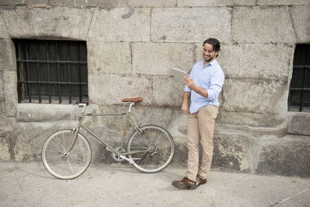 young happy attractive hispanic man in casual trendy clothes smiling cheerful using internet with digital tablet pad on vintage cool retro bicycle in modern stylish lifestyle urban style concept の写真素材